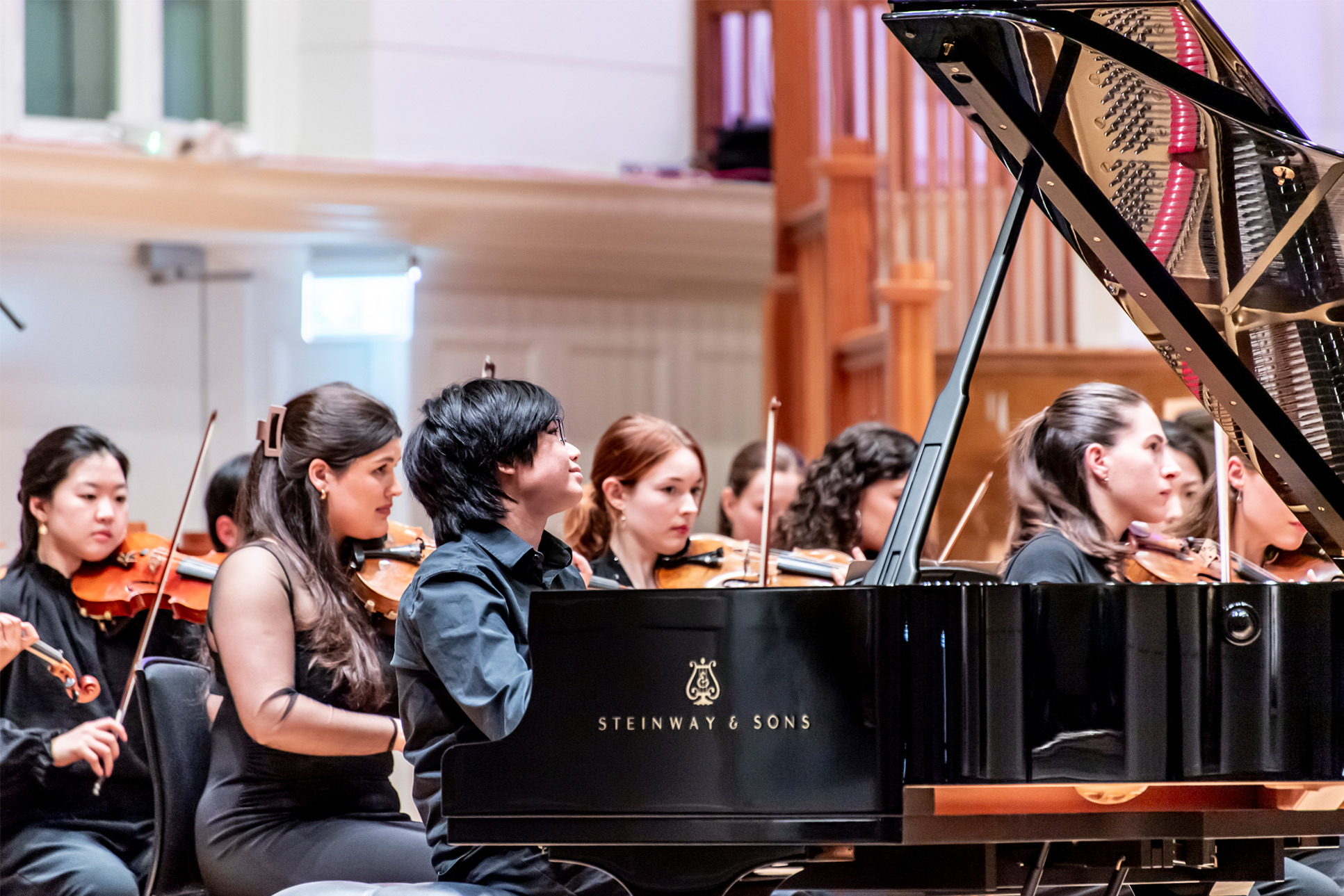 A student wearing smart black attire, playing the piano in an orchestra, with an orchestra being conducted in the background.